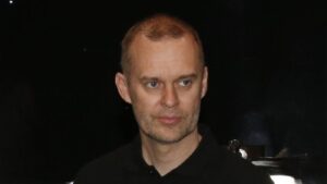Man with short light brown hair and stubble, wearing a black polo, looking to the right against a dark background.