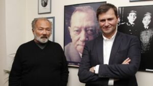 Two men stand beside large framed portraits in a gallery-like room, one with a gray beard wearing black, the other in a dark suit with arms crossed.