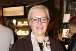 Smiling woman with short gray hair and glasses in a cosmetics store setting.