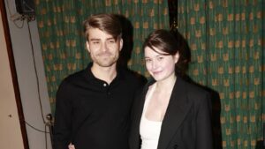 Man and woman posing for a photo at an indoor event, both smiling; man in a black polo, woman in a white top with a black blazer.