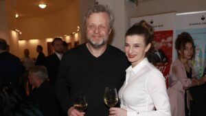 Smiling man with gray hair and stubble beside a woman in a white shirt, both holding white wine glasses at a reception.