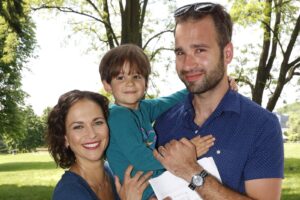 Smiling family of three outdoors in a park: mother, father, and young boy in the center.