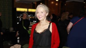 Smiling older woman with gray hair in a black lace top and red cardigan at a crowded indoor event.