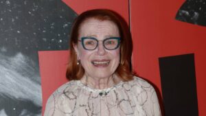 Smiling woman with blue glasses and red hair wearing a patterned blouse and pearl necklace at a red‑and‑black backdrop festival photo