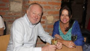 An older man and woman sit at a wooden table, smiling, each with a glass of white wine, against a brick wall.