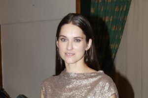 Portrait of a woman with long dark hair and hoop earrings, wearing a sparkling rose-gold top, looking at the camera indoors.