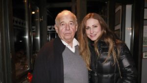 Older man and younger woman smiling together outdoors near a glass doorway.