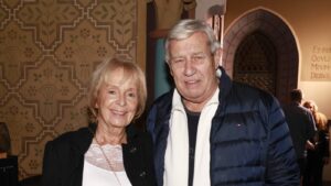 Smiling elderly couple posing together indoors, patterned wallpaper behind them and a decorative arch window to the right.