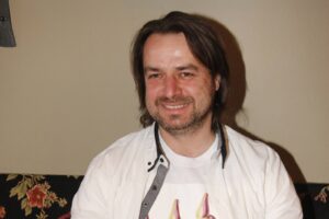 Portrait of a smiling man with dark hair and light stubble, wearing a white shirt indoors.