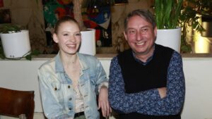 Two smiling friends sit side by side at a table in a cozy indoor space with plants in white pots behind them, posing for a photo.