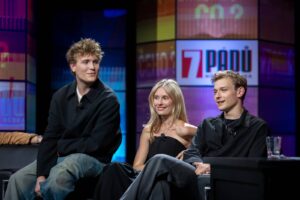 Three young adults sit on a TV talk-show set, each with a mic, framed by colorful panels and a visible '7 PADU' logo in the background.