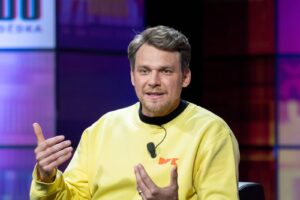 Man in a pale yellow sweatshirt speaks into a lapel microphone while gesturing with his hands in a studio set with purple lighting.