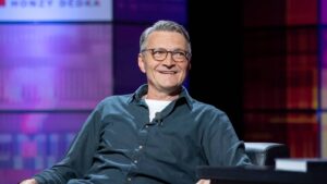 Smiling man with gray hair and glasses sits on a stage, wearing a dark blue button-down shirt with a lavalier mic clipped to it.