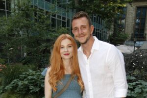 Couple posing outdoors in a garden, smiling. Woman with long red hair in a blue dress, man in a white shirtVisible behind glass building.