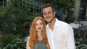 Couple posing outdoors in a garden, smiling. Woman with long red hair in a blue dress, man in a white shirtVisible behind glass building.