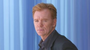 Portrait of a man with reddish-blonde hair in a dark suit standing against a blue vertical-panel backdrop.