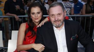 Woman in a red sequined dress sits beside a man in a dark suit, arm around his shoulder, posing for a photo at a crowded event behind a barricade