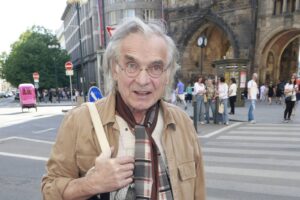 Older man with gray hair and round glasses standing at a busy city crosswalk, beige jacket and scarf visible in front of a historic building.