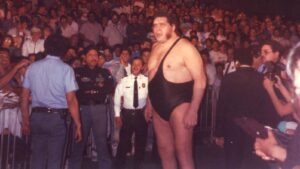 A large wrestler in a black one‑shoulder singlet stands among fans and photographers at a wrestling event.