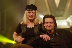 Two performers pose on stage: a blonde woman in a black outfit and beret beside a smiling man with long hair, rope in the foreground.