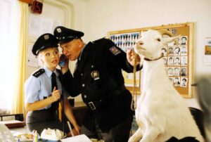 Two police officers in a cluttered office, one on the phone, with a white llama beside them holding a rope around its neck like a prop.