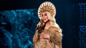 Singer in a gold sequin gown with a large ornate sunburst headdress singing into a microphone on stage.