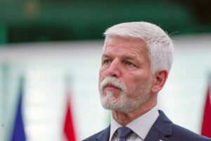 Portrait of an older man with white hair and beard in a dark suit, looking thoughtful, with flags in the blurred background.
