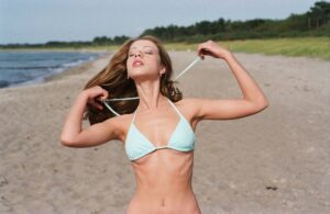 Woman in a light blue bikini on a sandy beach, pulling on her bikini straps with eyes closed and wind in her hair, shoreline in background