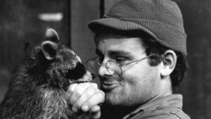 Man wearing a cap and glasses leans in to nuzzle a furry animal, sharing a tender moment.