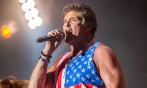 Male singer wearing a sleeveless American flag shirt, singing into a handheld microphone on stage.