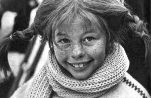 A smiling young girl with freckles wearing a knit scarf and braided pigtails in black-and-white photograph.