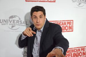 Young man in a dark blazer points at the camera in front of a backdrop that reads Universal 100th Anniversary and American Pie Reunion.