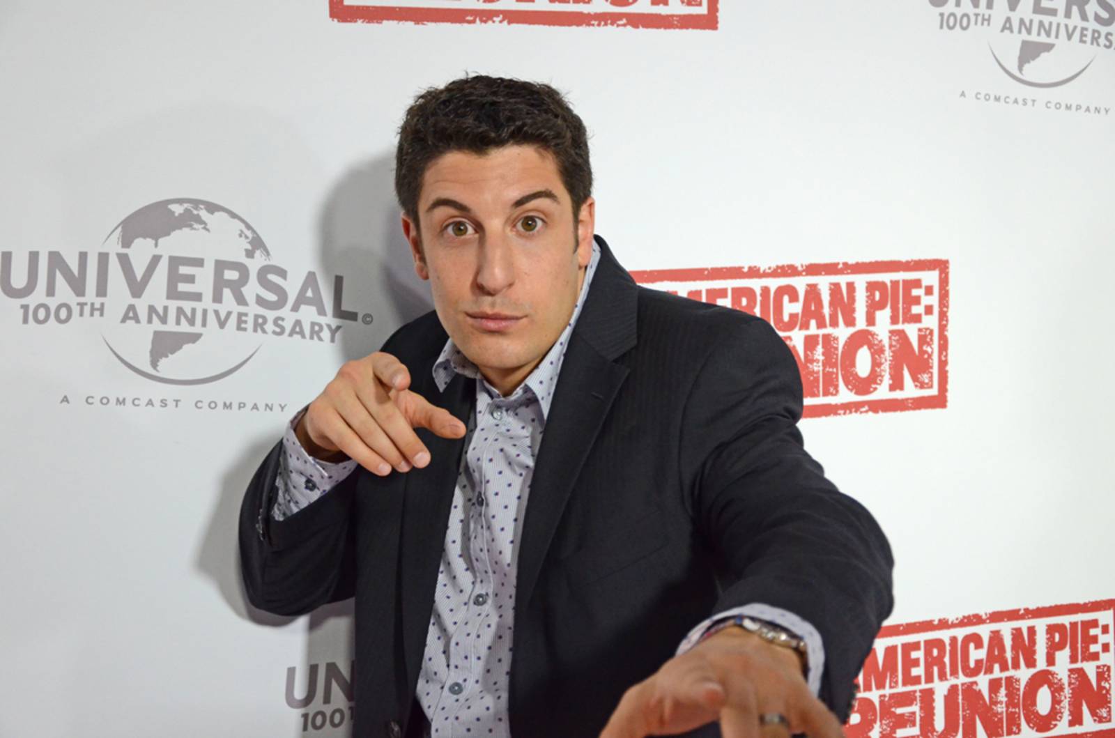 Young man in a dark blazer points at the camera in front of a backdrop that reads Universal 100th Anniversary and American Pie Reunion.