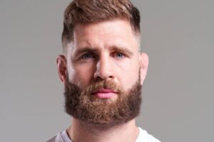 Close-up portrait of a man with a full brown beard and short hair against a gray background.
