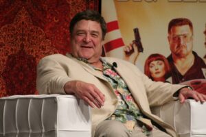 Smiling man in a beige suit and tropical shirt seated on a white couch on stage with a patterned backdrop.
