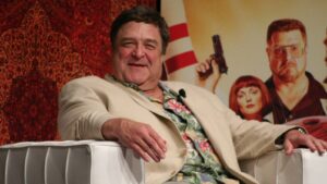 Smiling man in a beige suit and tropical shirt seated on a white couch on stage with a patterned backdrop.