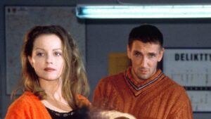 A woman with long blond hair in an orange cardigan stands in the foreground while a man in an orange knit sweater looks on in a dim room with a blurred sign in the background.