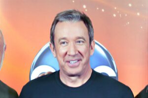 Smiling man with short gray hair wearing a black shirt against an orange backdrop.