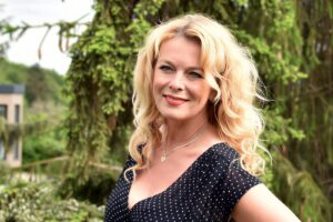 Portrait of a smiling blonde woman in a black polka dot dress, outdoors with greenery in the background, wearing a silver necklace.
