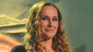 Close-up of a smiling woman with long, curly hair on stage under warm lights in a dark outfit.