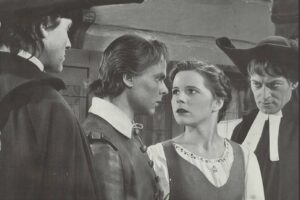 Four adults in period costumes stand close, facing each other in a tense conversation indoors.