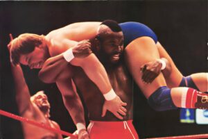 Two male wrestlers in a match: one carries the other over his shoulder as others watch.
