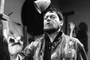Black-and-white photo of a bearded painter in a paint-splattered jacket, cigarette in mouth, standing at an easel in a cluttered studio.