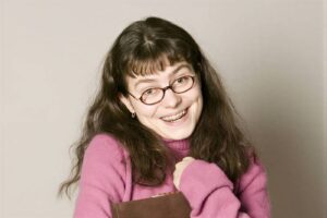 Smiling girl with braces and round glasses, wearing a pink sweater and holding a brown notebook.