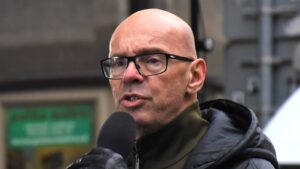 Bald man wearing glasses speaks into a handheld microphone outdoors, wearing a dark jacket with a hooded coat visible behind him—public interview setting.