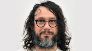 Portrait of a man with shoulder-length dark hair, round black glasses, and a gray beard, looking at the camera against a light background.