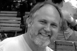 Close-up of a smiling man with a beard, outdoors with wooden benches in the background (black and white).