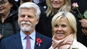 Smiling older man in a navy suit with a red poppy pin and a blonde woman wearing a pink scarf in a group photo at a ceremony.