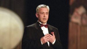 Older man in a black tuxedo and red bow tie speaking on stage, hands pressed to chest.