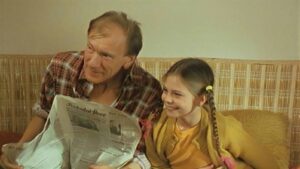 An older man and a young girl sit on a striped couch, smiling while reading a newspaper together.